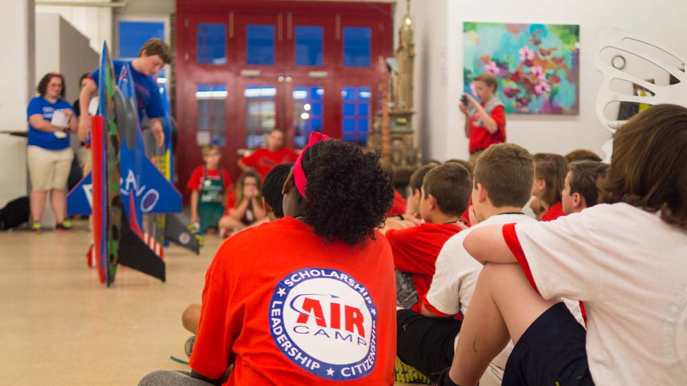 air camp students in a circle watching other student displaying their painted plane models